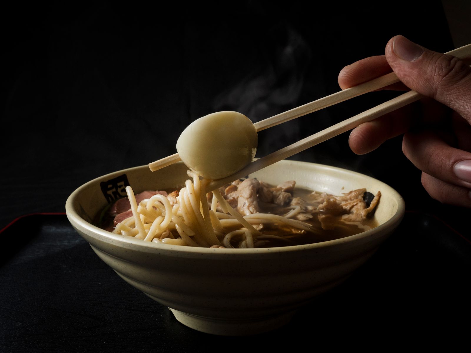 Steam rising off a hot ramen bowl as chopsticks lift a marinated egg